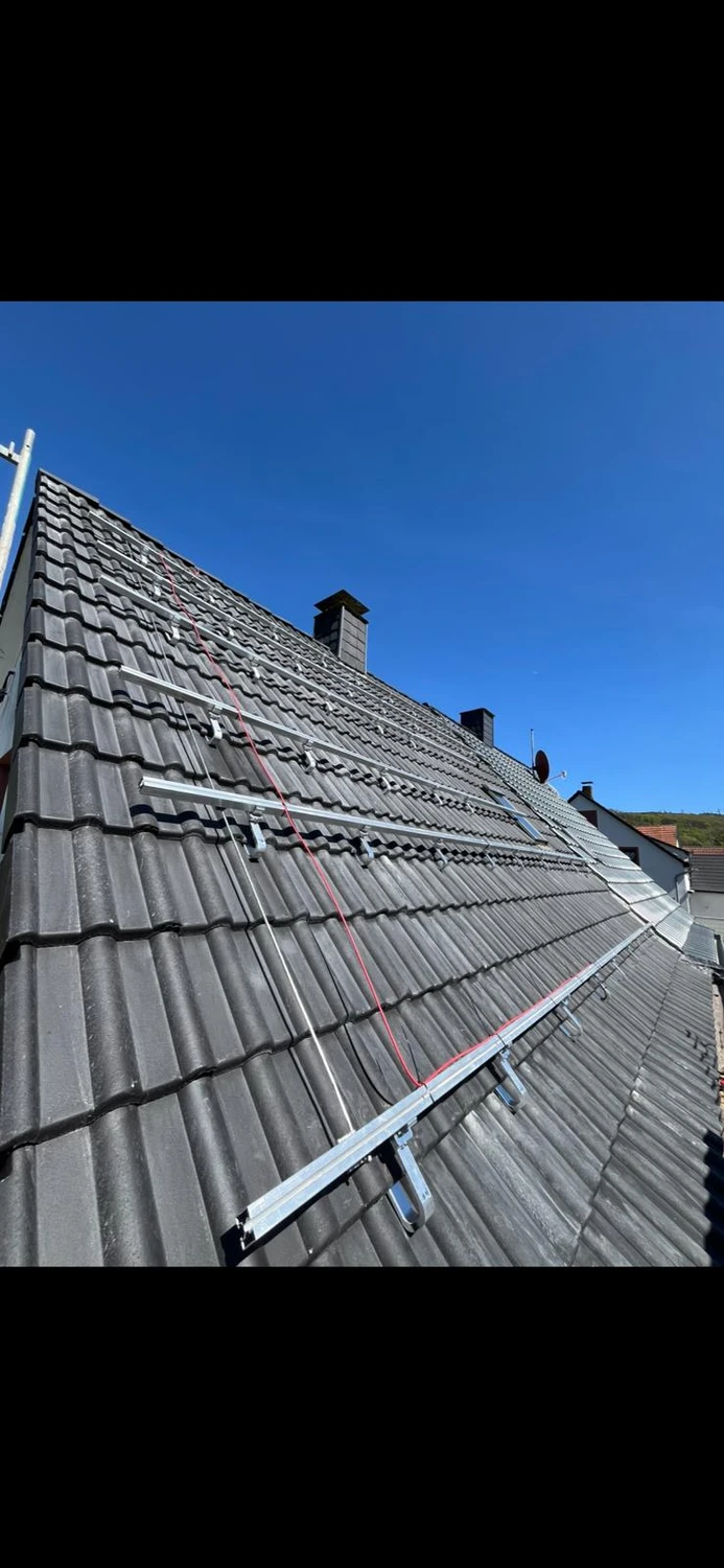 Spanndecken Grunau - Sanierung, Renovierung & Innenausbau - Nahaufnahme eines Schrägdachs mit dunkelgrauen Ziegeln, auf dem horizontale Aluminiumschienen und Führungsschienen für die Installation von Solarmodulen unter klarem, blauem Himmel zu sehen sind.