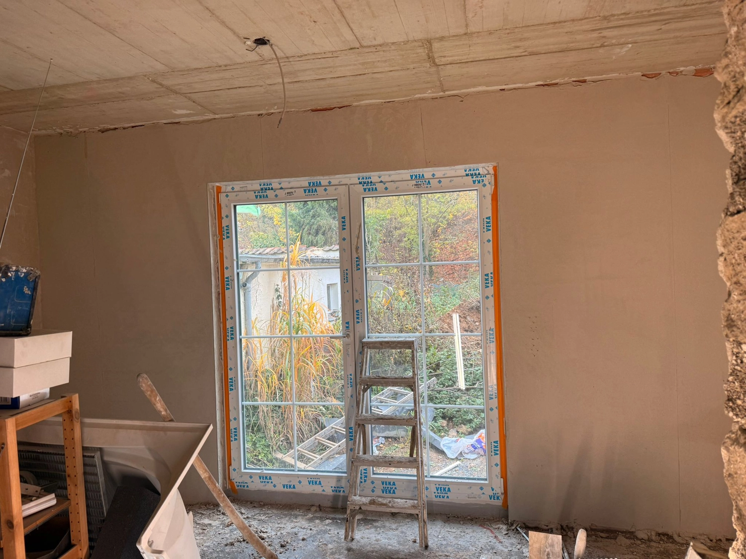 Spanndecken Grunau - Sanierung, Renovierung & Innenausbau - Ein Innenraum im Umbau. Die Wand ist frisch verputzt und grau. Eine Doppeltür aus Glas und eine Metallleiter sind eingebaut.